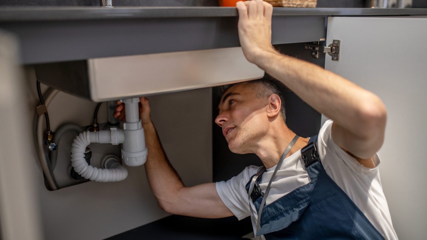 Plumber, repair. Experienced attentive middle-aged man carefully examining bottom of kitchen sink touching pipe with his hand diagnosing problem